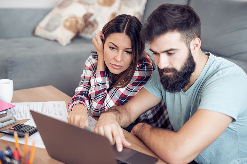 A couple sits together at a table, looking concerned while reviewing documents and using a laptop. Papers, a calculator, and a mug are on the table, suggesting they are managing finances or budgeting at home.