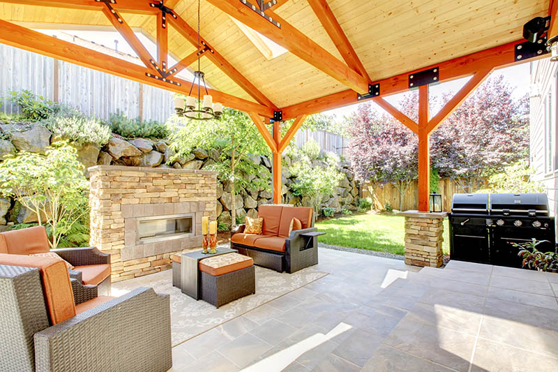 Covered patio with wooden beams, wicker furniture, a stone fireplace, and a barbecue grill. Lush greenery and shrubs surround the outdoor space, creating a cozy and inviting backyard setting.
