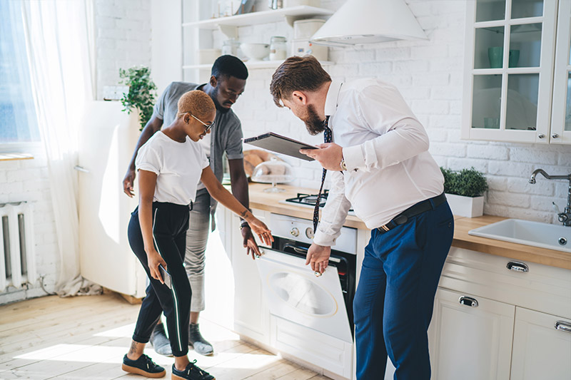 A man in a suit shows a couple an oven in a modern white kitchen. The couple looks closely at the appliance while the man holds a clipboard and gestures toward the open oven door.