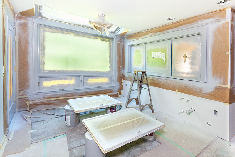 A room prepared for painting with plastic coverings on the floor and windows, two cabinet doors on sawhorses, a step ladder, and paint supplies scattered around. The walls and woodwork are primed and ready for paint.