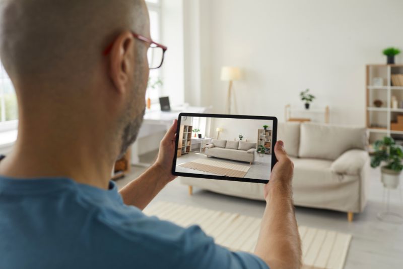 A person holding a tablet takes a photo of a modern living room with a beige sofa, a rug, and plants. The room appears bright and neatly decorated with natural light from the windows.