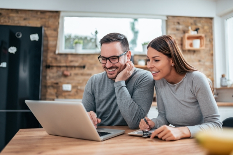 A smiling couple sits at a kitchen table looking at a laptop. The man is wearing glasses and both are dressed casually. The woman holds a credit card and a notepad. The kitchen has a brick wall and a window in the background.
