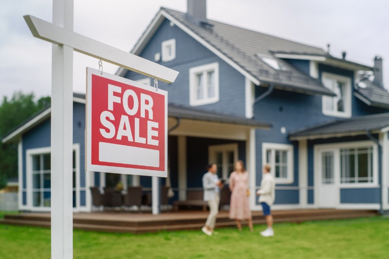 A red and white For Sale sign is in the foreground, with a modern blue house and three people talking on the front lawn in the background.