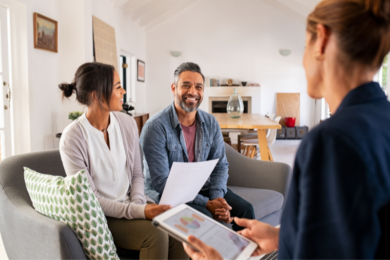 A smiling couple sits on a couch holding paperwork, facing a professional woman with a tablet, having a discussion in a bright, modern living room.