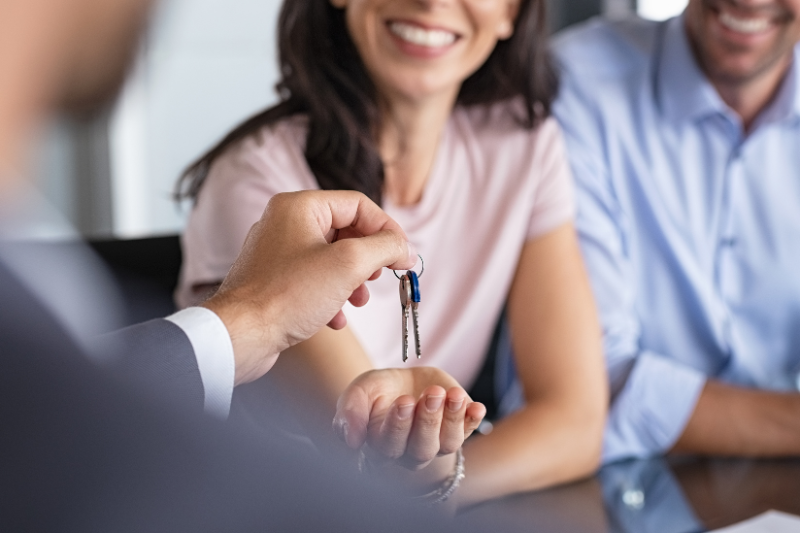 A person hands a set of keys to a smiling woman, who is reaching out her hand, while another person sits beside her. The scene suggests the purchase or rental of a new home.