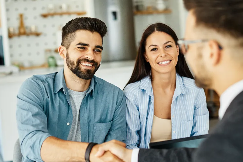 A smiling man and woman sit together at a table, shaking hands with another person in business attire across from them in a bright, modern office setting.